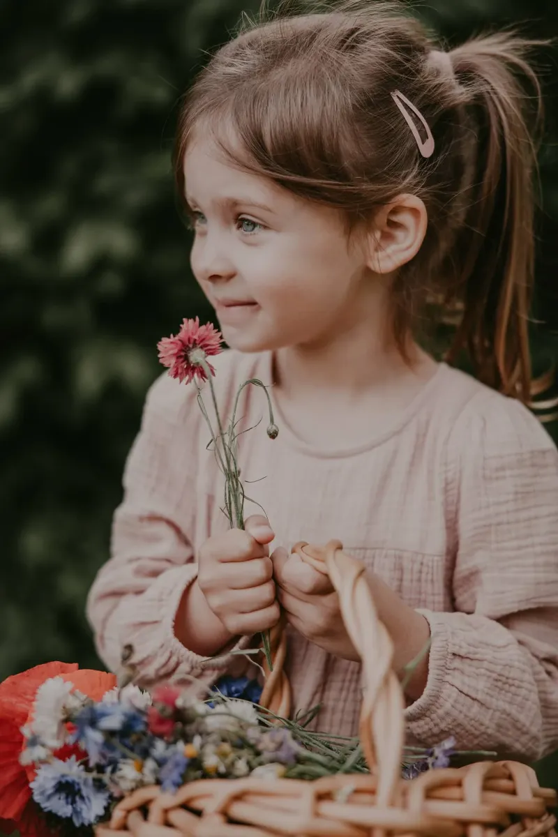 Mädchen in rosa Kleidung hält eine Blume, Korb mit Wiesenblumen im Vordergrund